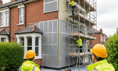 A UK terraced house being retrofitted under PAS 2035 with workers in hi vis jackets installing external wall insulation and a clipboard of technical drawings in the foreground.