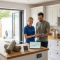 A qualified domestic energy assessor reviewing home energy performance with a UK homeowner in a bright kitchen, with insulation, smart controls, solar equipment and an air source heat pump visible.
