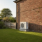 A modern white air source heat pump outdoor unit installed against the rear brick wall of a UK semi detached home, sitting on a low concrete plinth in a tidy back garden, demonstrating compliant permitted development positioning.