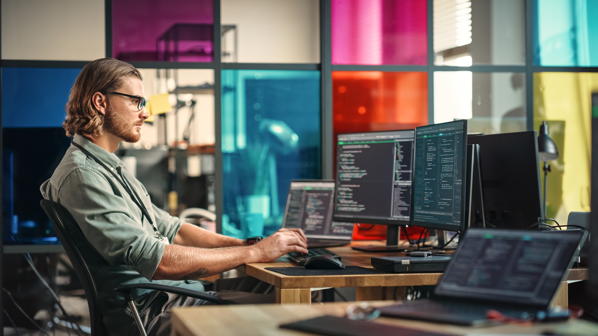 A man with glasses types code on multiple monitors in a modern office with colorful glass panels.