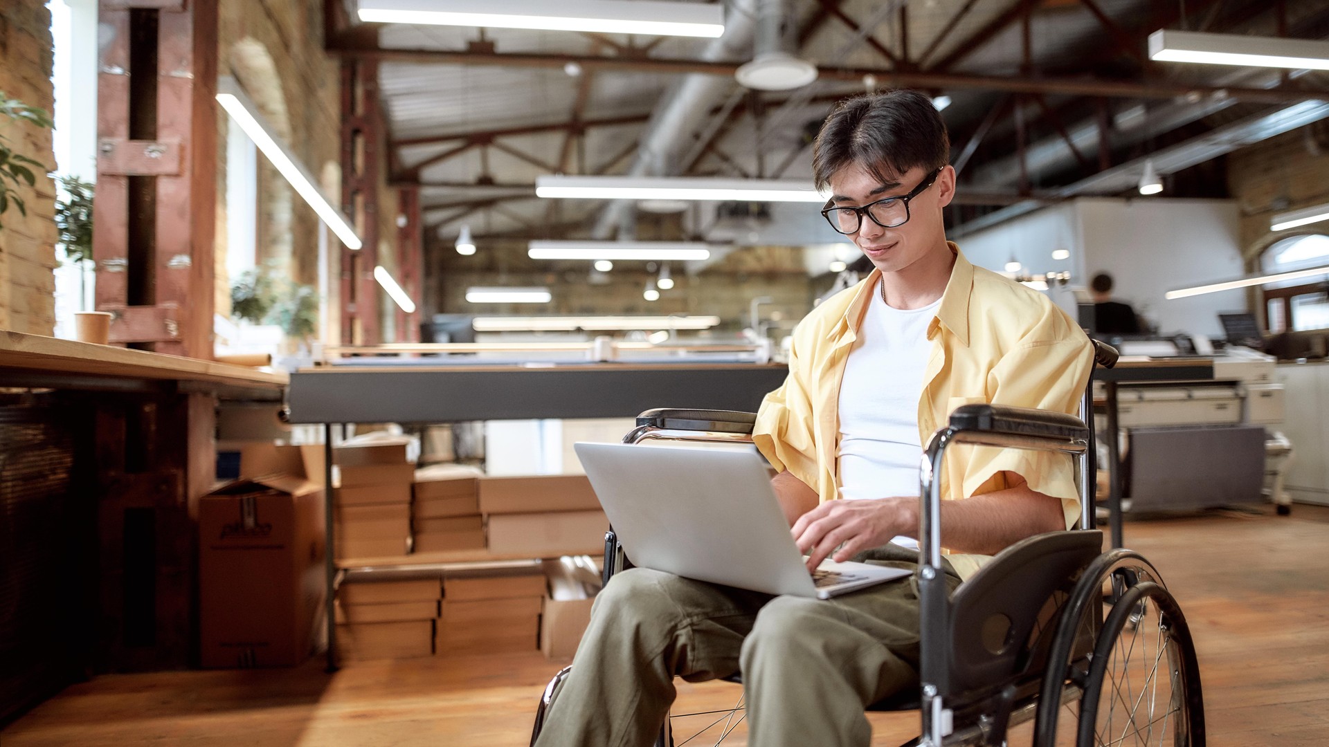 A young man in a wheelchair smiles as he types on a laptop in a modern office.