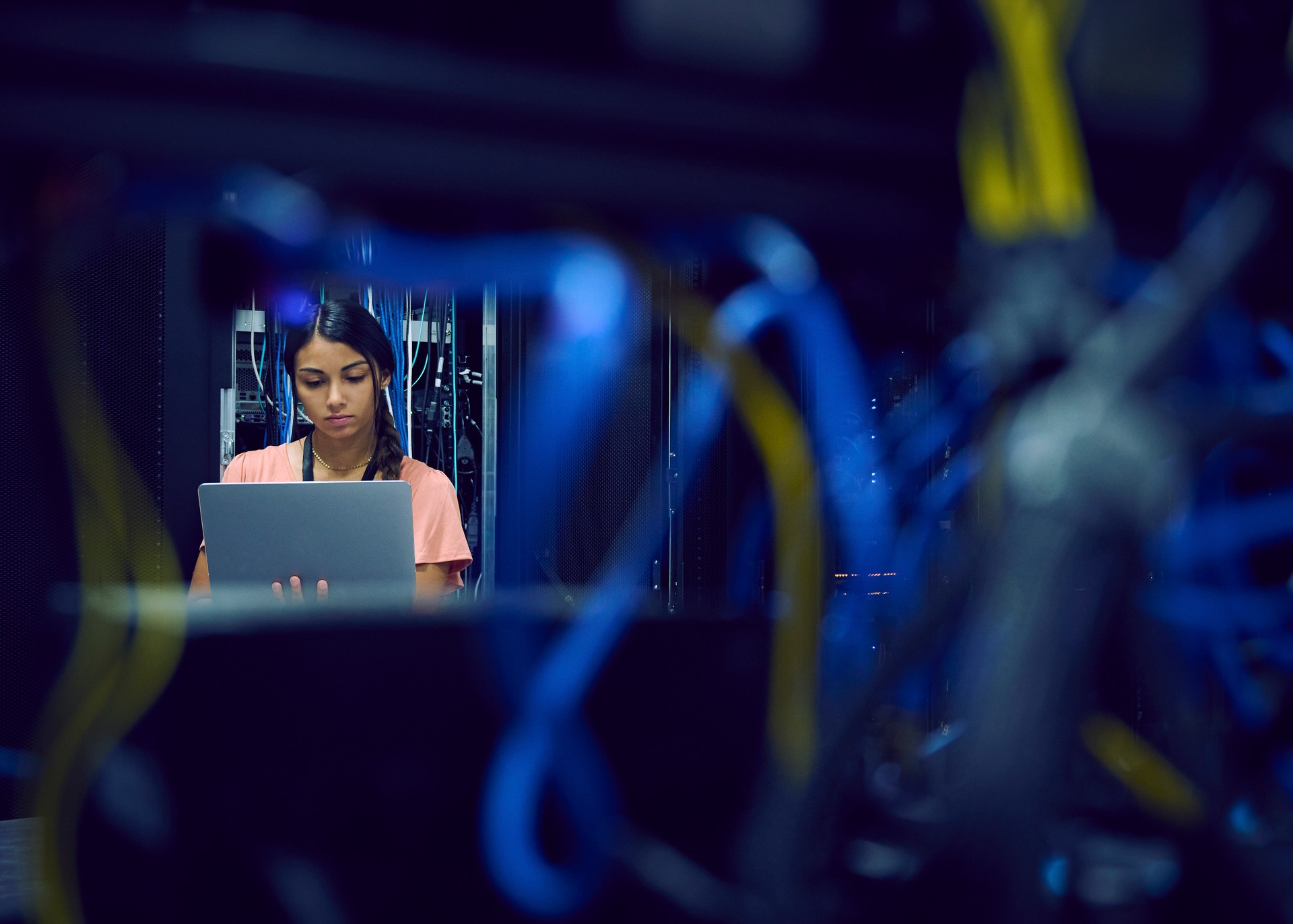 A woman works on a laptop in a data center, with blurred network cables in the foreground.
