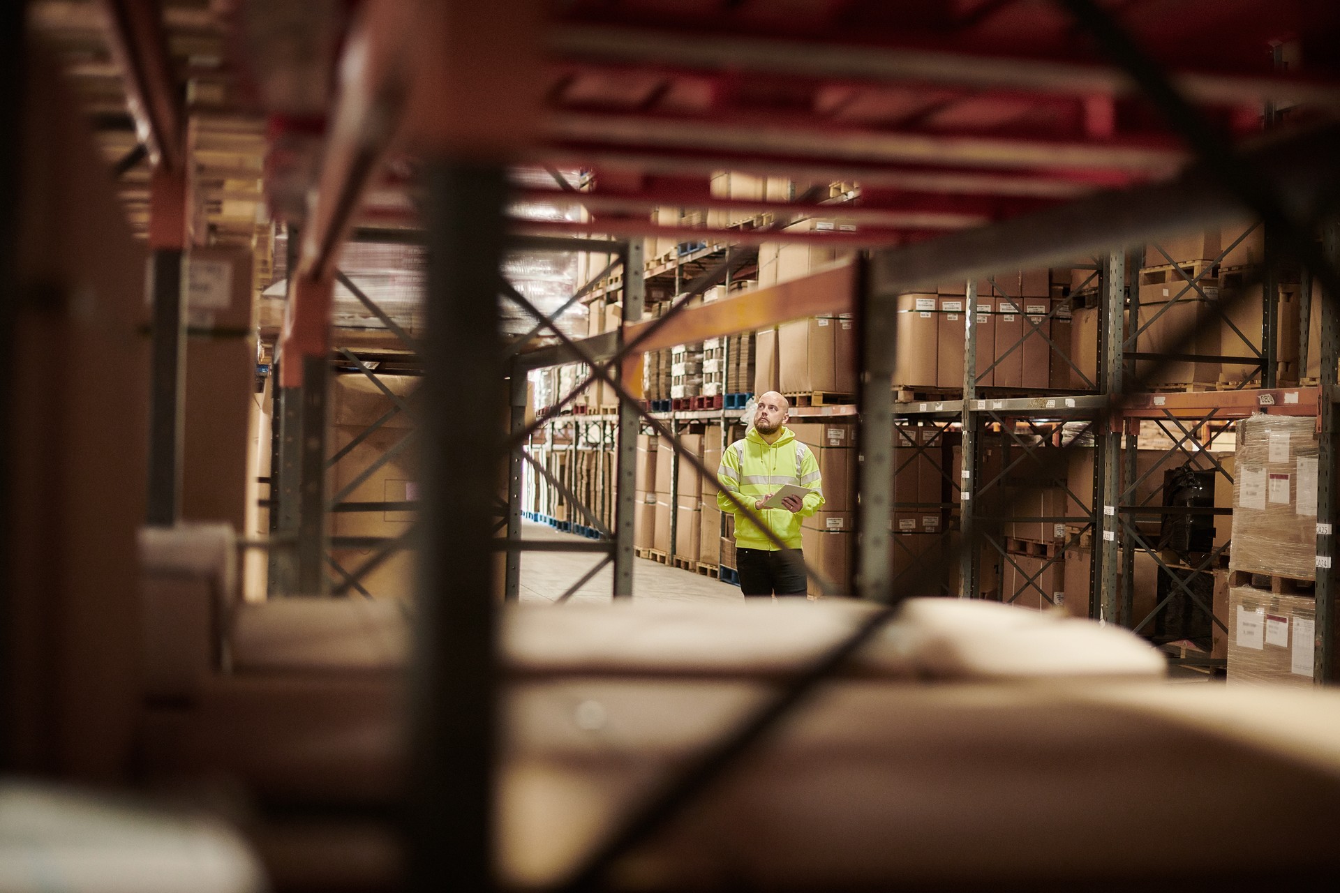 A man in a reflective vest holds a tablet while standing in a large warehouse aisle filled with boxes on shelves.
