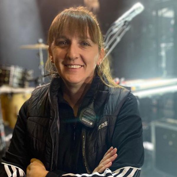 Photo de Laetitia Leroy bras croisés, souriante sur une scène, on peut apercevoir des cases protégreant les instruments et une batterie