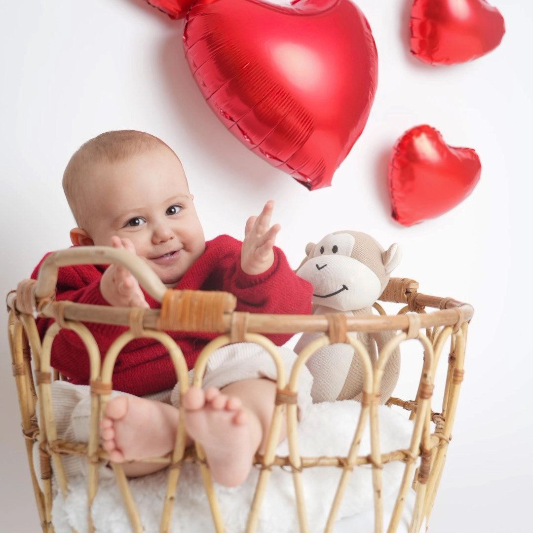 Ein lachendes Baby sitzt mit Kuscheltier im Korb unter roten Herzballons.