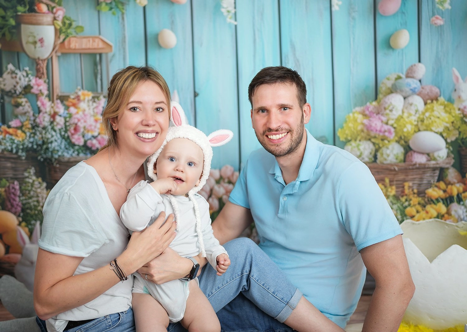 Zwei Erwachsene halten lächelnd ein Baby mit Hasenmütze zwischen bunten Blumen.