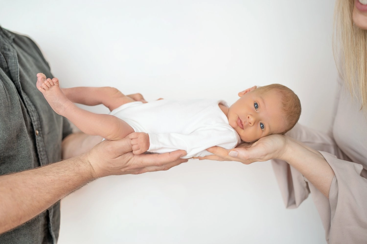 Ein waches Baby in weißer Kleidung liegt auf vier erwachsenen Händen.