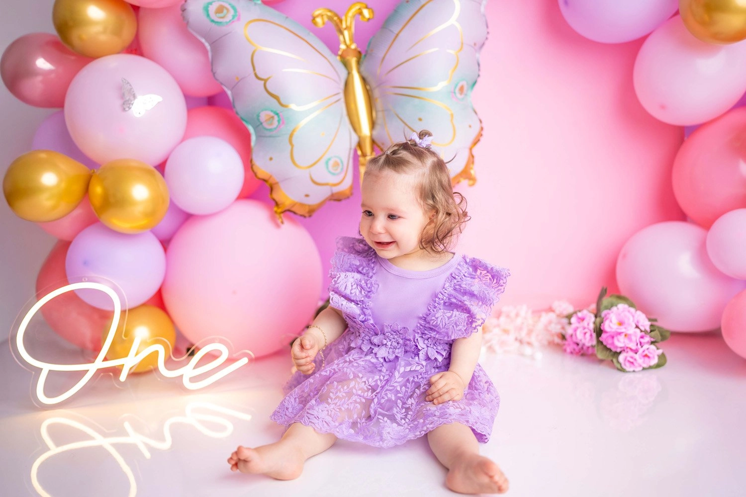 Ein Kind im lilafarbenen Kleid sitzt lächelnd vor rosa Ballondekoration.