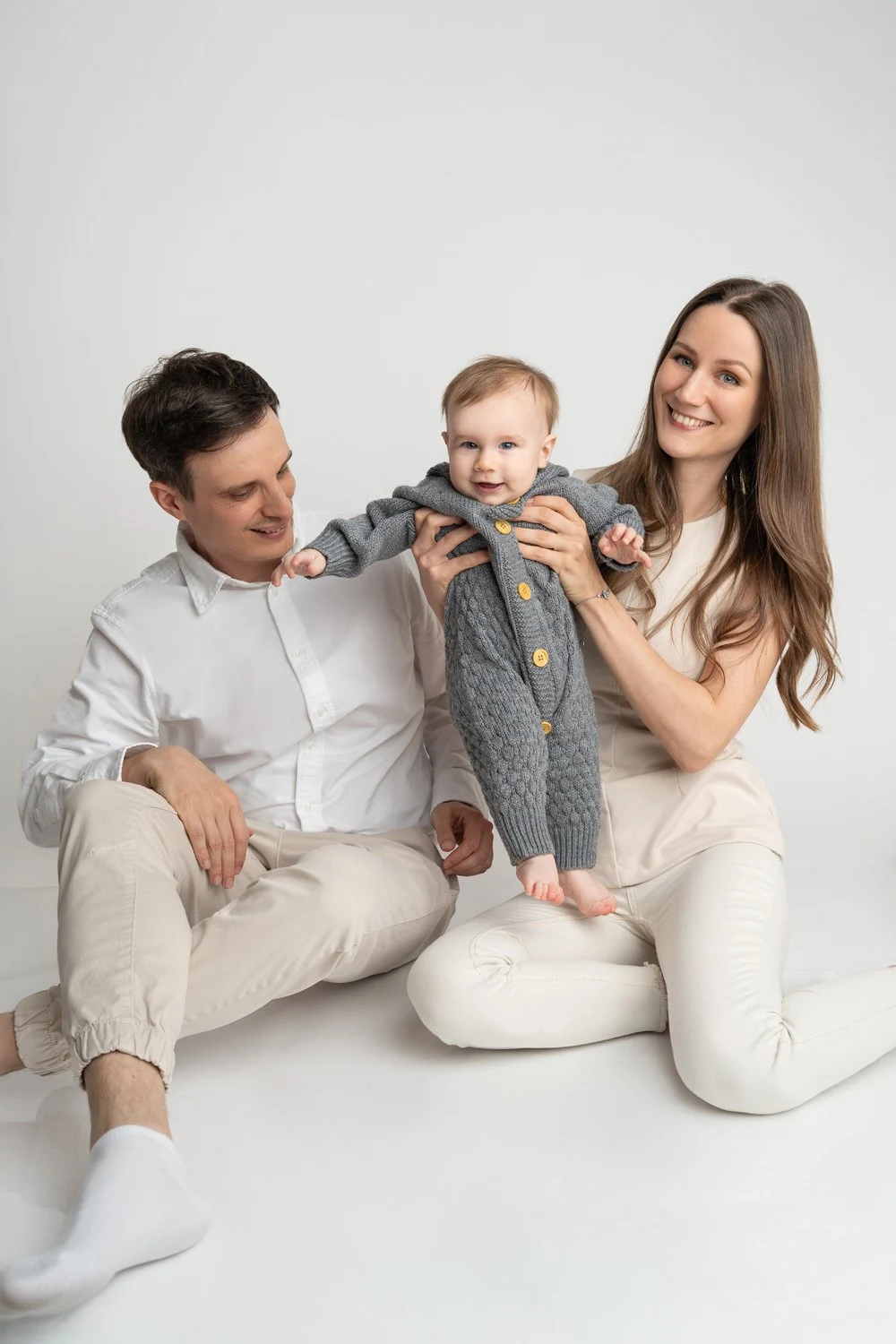Zwei Erwachsene sitzen in heller Kleidung mit einem Baby im Studio.