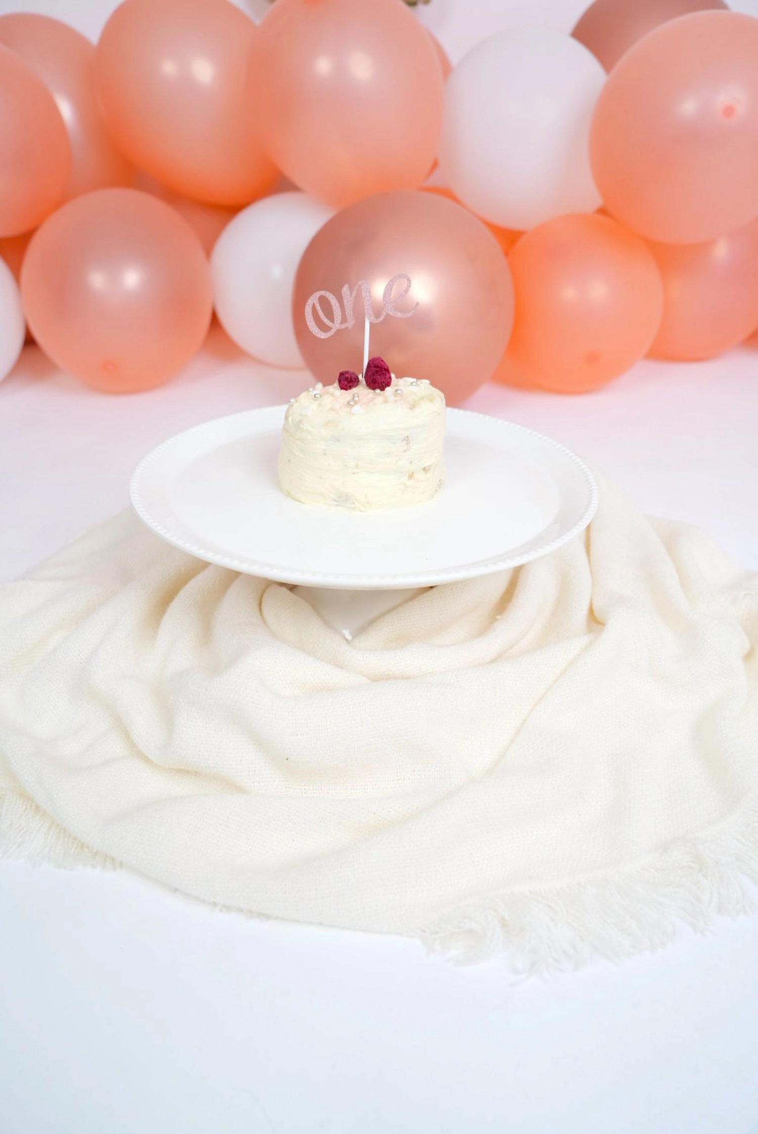 Eine kleine Torte mit One-Dekoration steht vor pfirsichfarbenen Ballons.