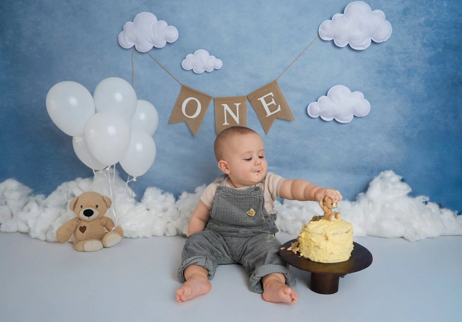 Ein Baby sitzt neben einer Torte vor blauer Wolkendekoration.