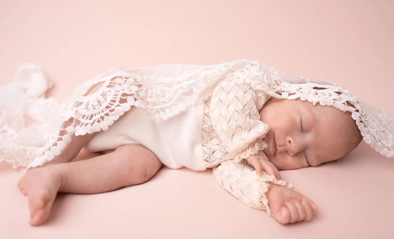 Ein schlafendes Baby mit Spitzenhaube liegt seitlich auf rosa Stoff.