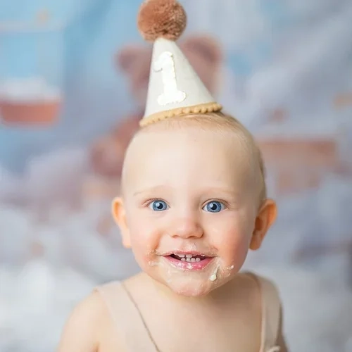 Ein lachendes Baby trägt einen kleinen Geburtstagshut und etwas Tortencreme.