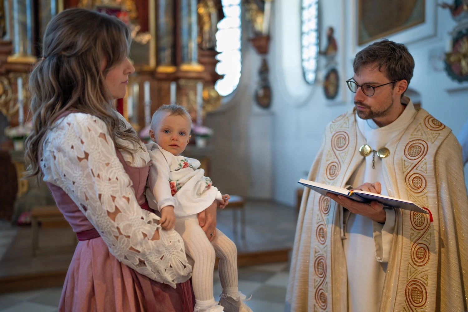 Eine Frau hält ein Baby, während ein Pfarrer aus einem Buch liest.