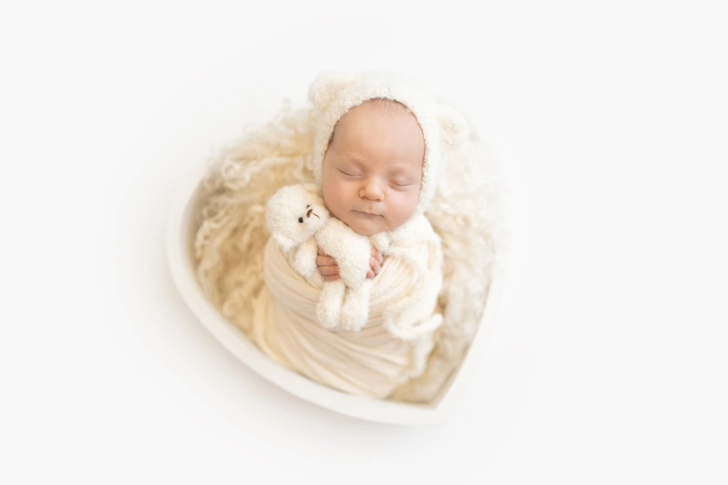 Ein schlafendes Baby liegt mit kleinem Kuscheltier in einem hellen Herzkörbchen.