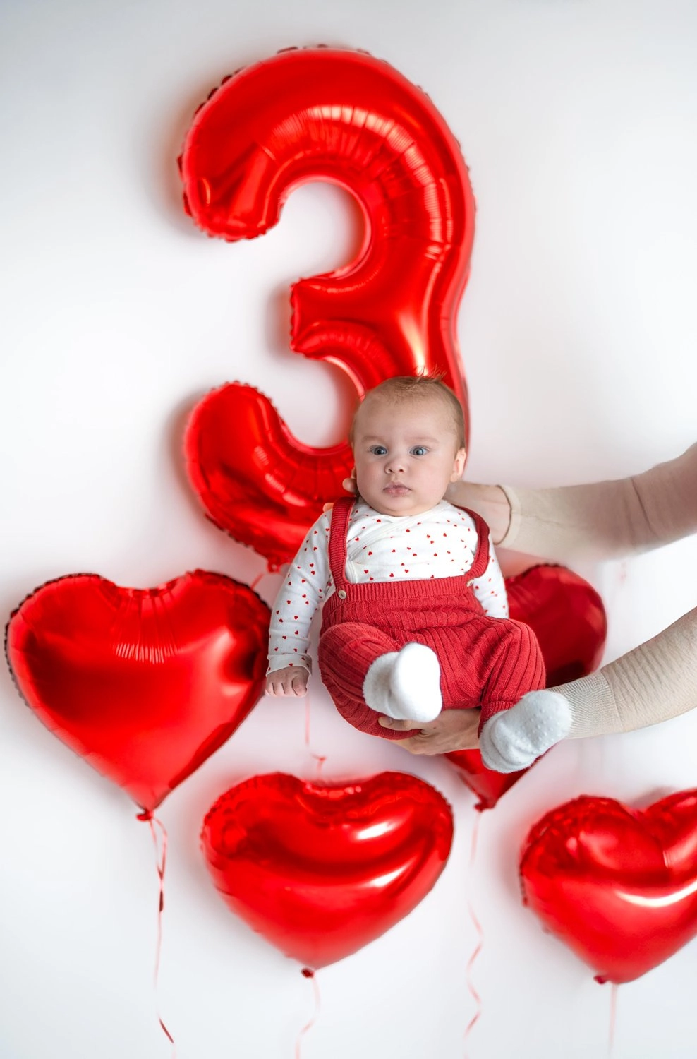 Ein Baby wird zwischen roten Herzballons und einer großen Drei gehalten.