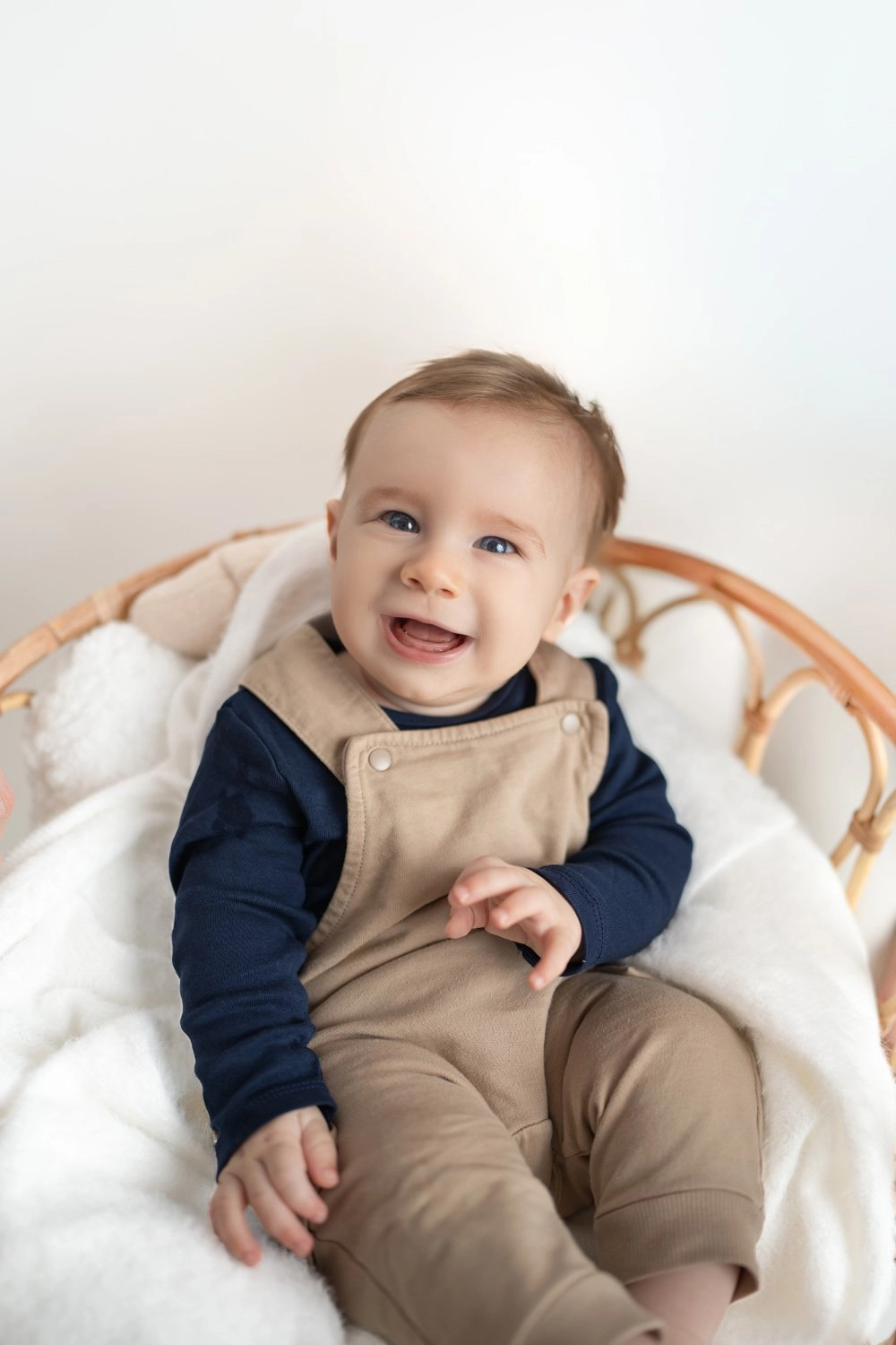 Ein lachendes Baby in beigem Outfit sitzt in einem Rattansessel.