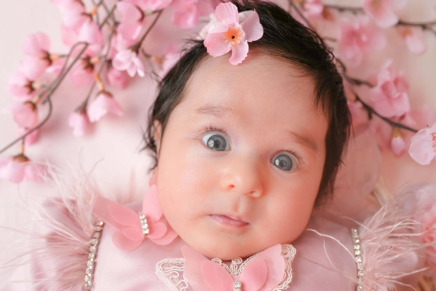 Ein waches Baby mit dunklen Haaren liegt unter rosa Blüten.
