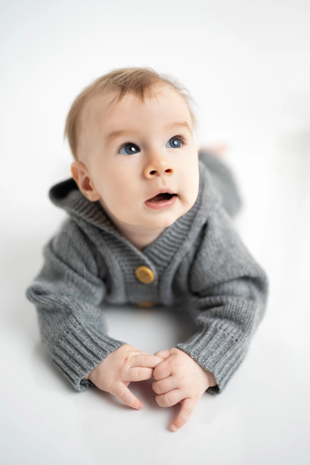 Ein Baby im grauen Strickpullover liegt und blickt neugierig nach oben.