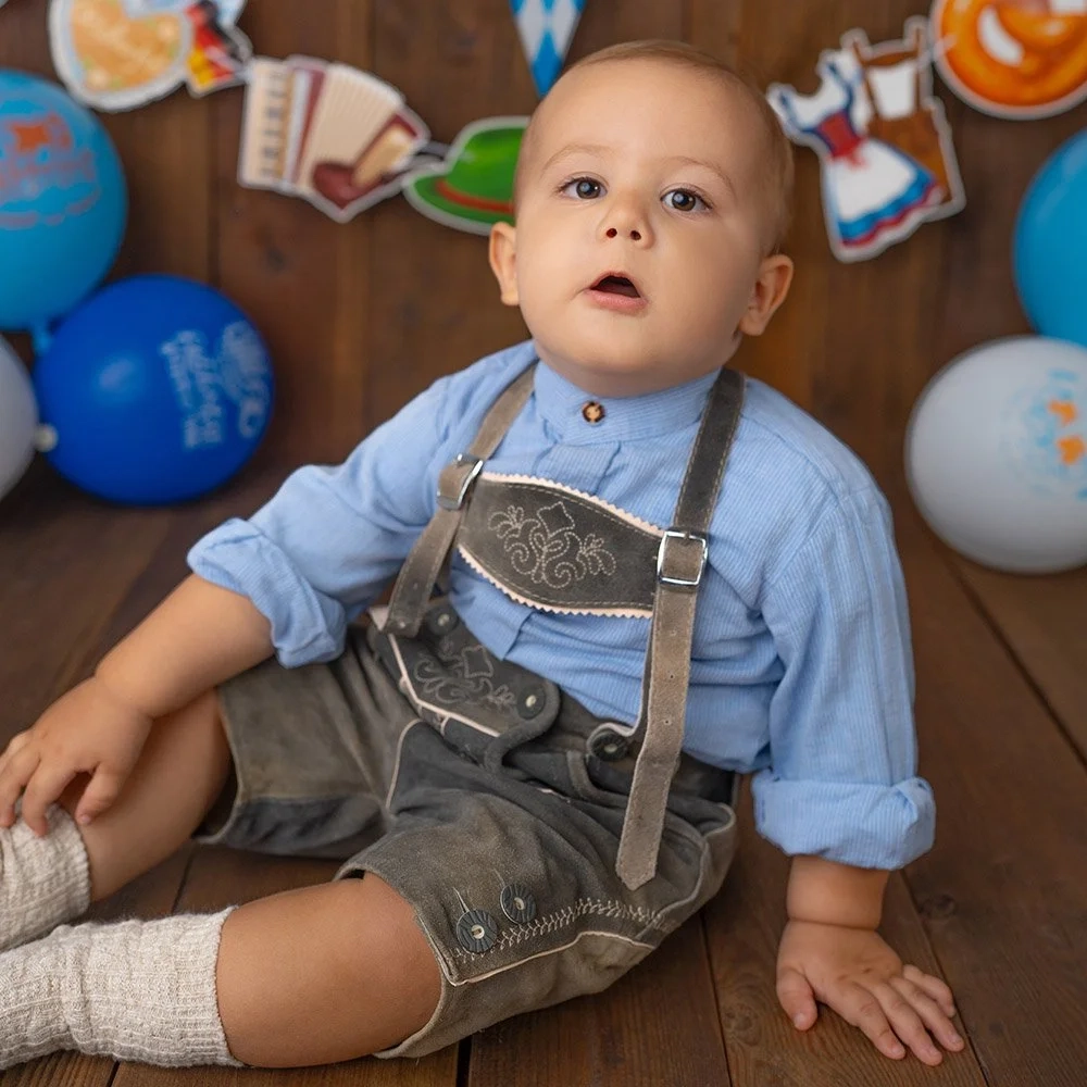 Ein Baby mit blauer Bluse und Lederhose sitzt auf Holzboden.