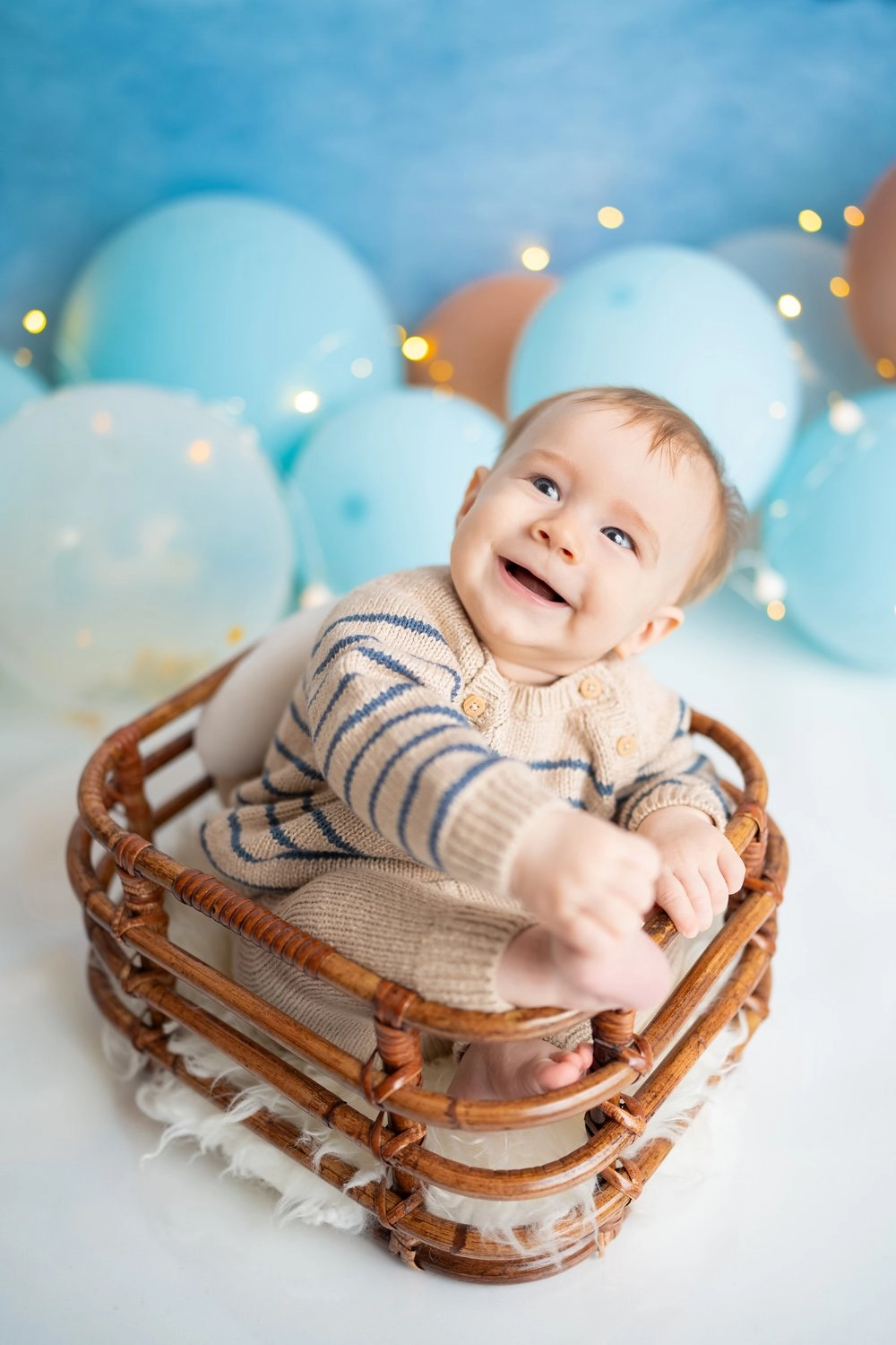 Ein lachendes Baby im Streifenpullover sitzt zwischen blauen Ballons im Körbchen.