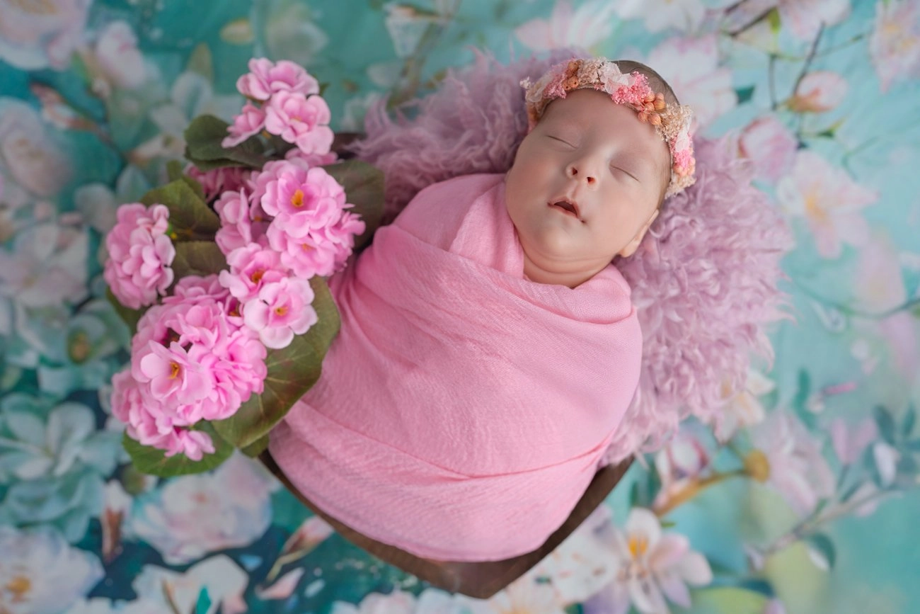 Ein schlafendes Baby liegt in rosa Stoff zwischen zarten Blüten.