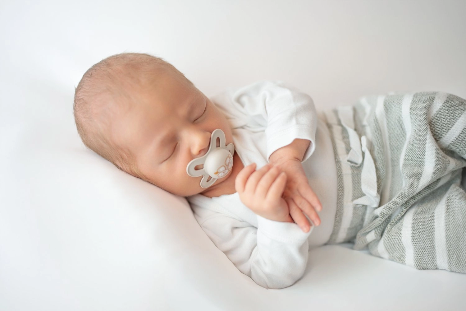 Ein schlafendes Baby mit Schnuller liegt in gestreifter Hose auf hellem Stoff.