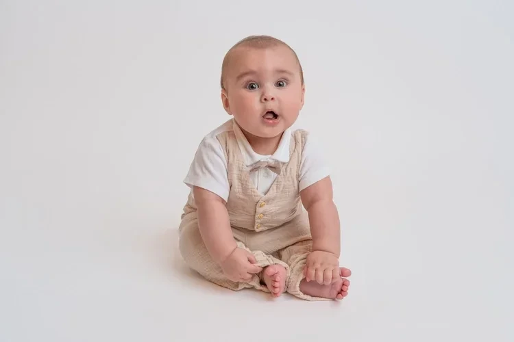 Ein Baby in beiger Latzhose sitzt aufmerksam auf hellem Boden.
