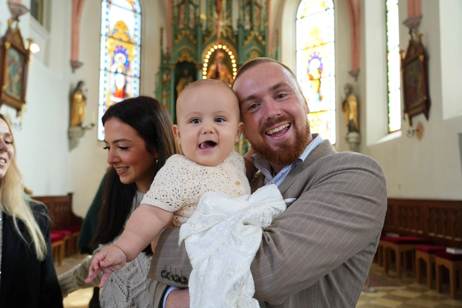 Zwei lächelnde Erwachsene halten ein Baby im weißen Kleid in einer Kirche.