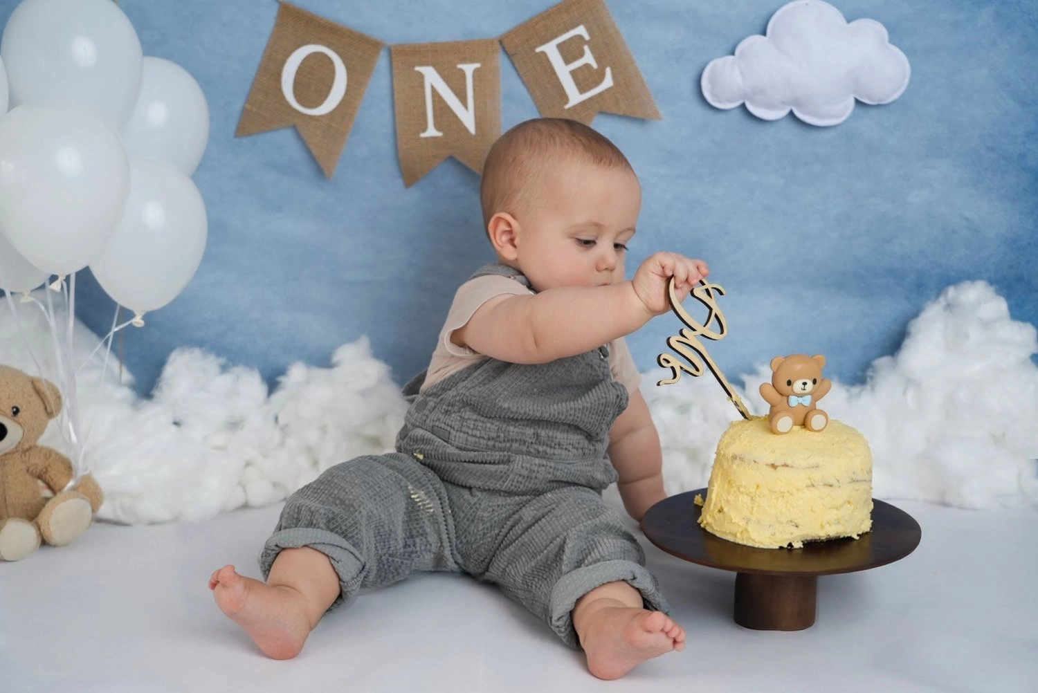 Ein sitzendes Baby untersucht seine Torte vor einer blauen Wolkenkulisse.