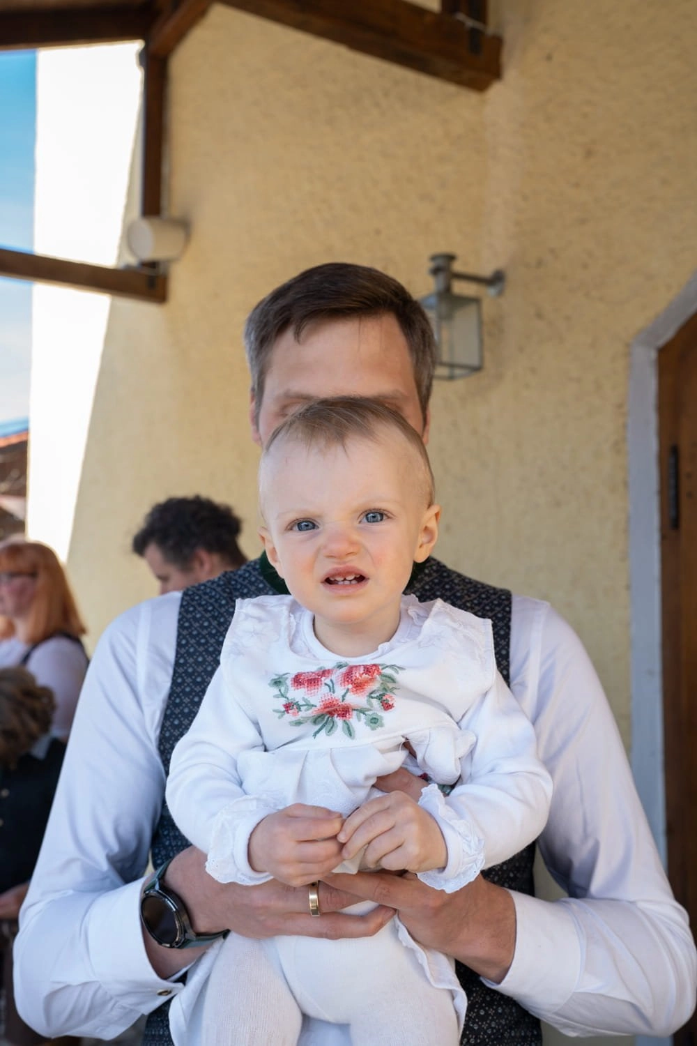 Ein Baby im weißen Kleid wird von einem Mann gehalten.