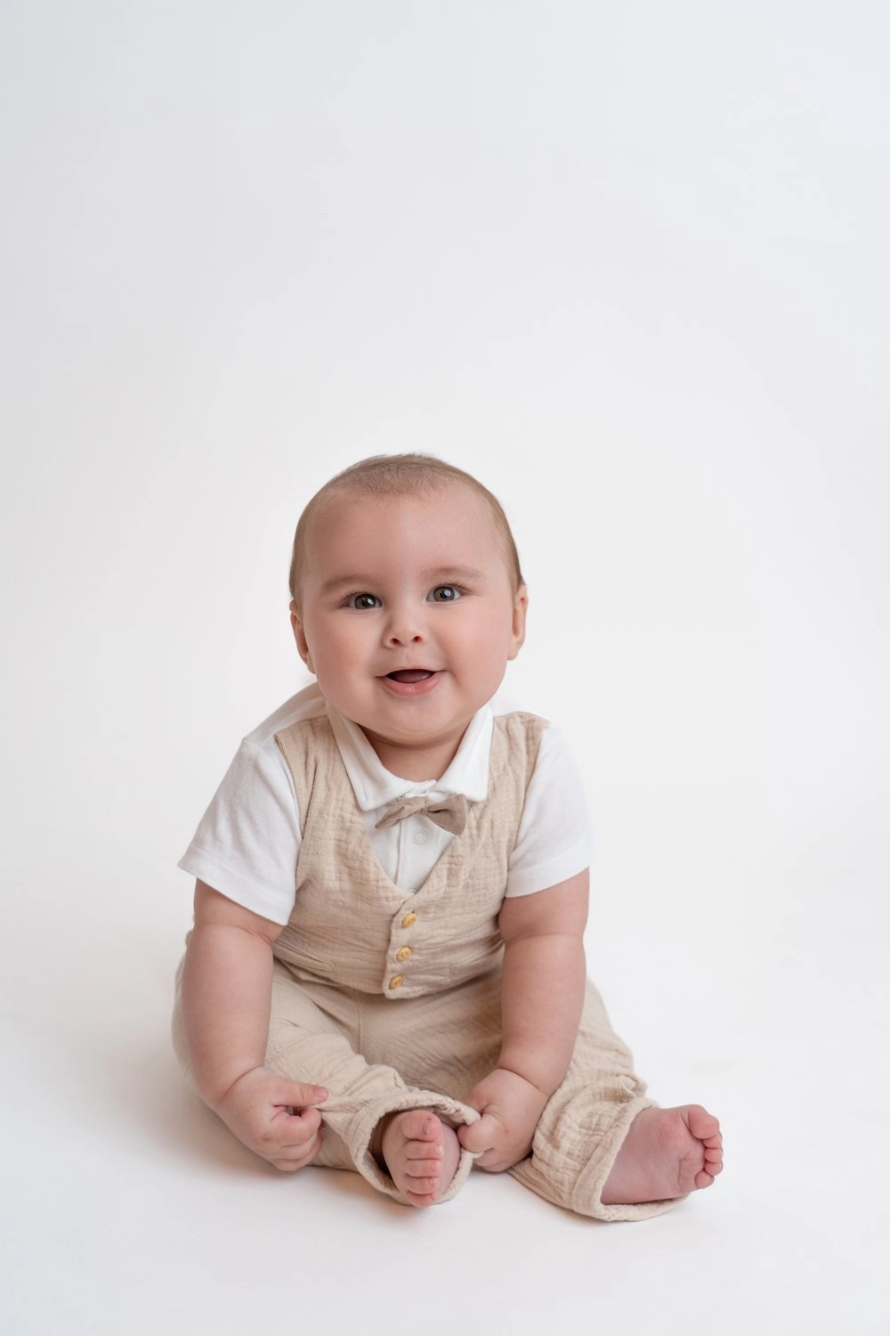 Ein Baby in beiger Latzhose sitzt lächelnd auf hellem Untergrund.