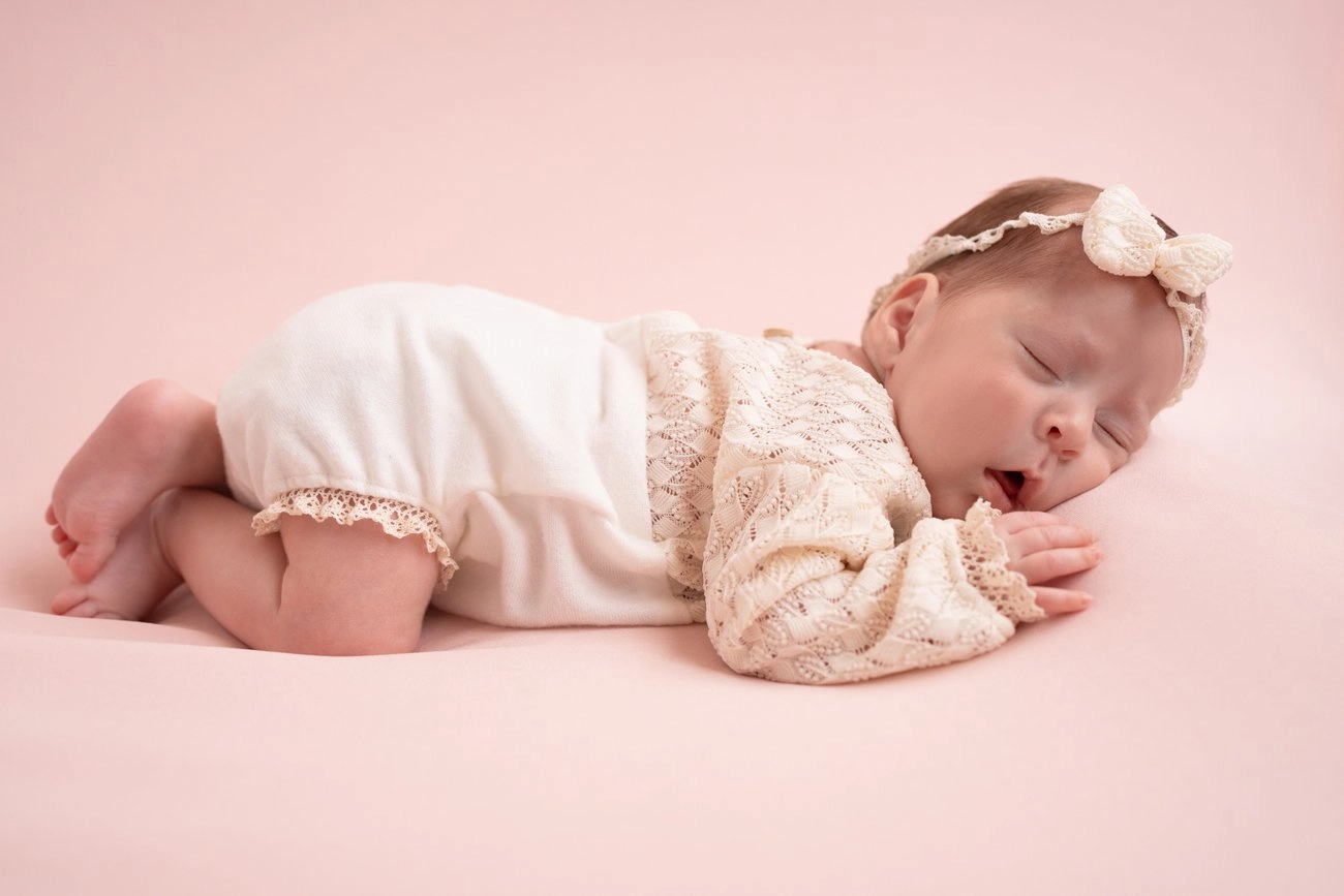 Ein Baby in heller Spitzenkleidung liegt gähnend auf rosa Untergrund.
