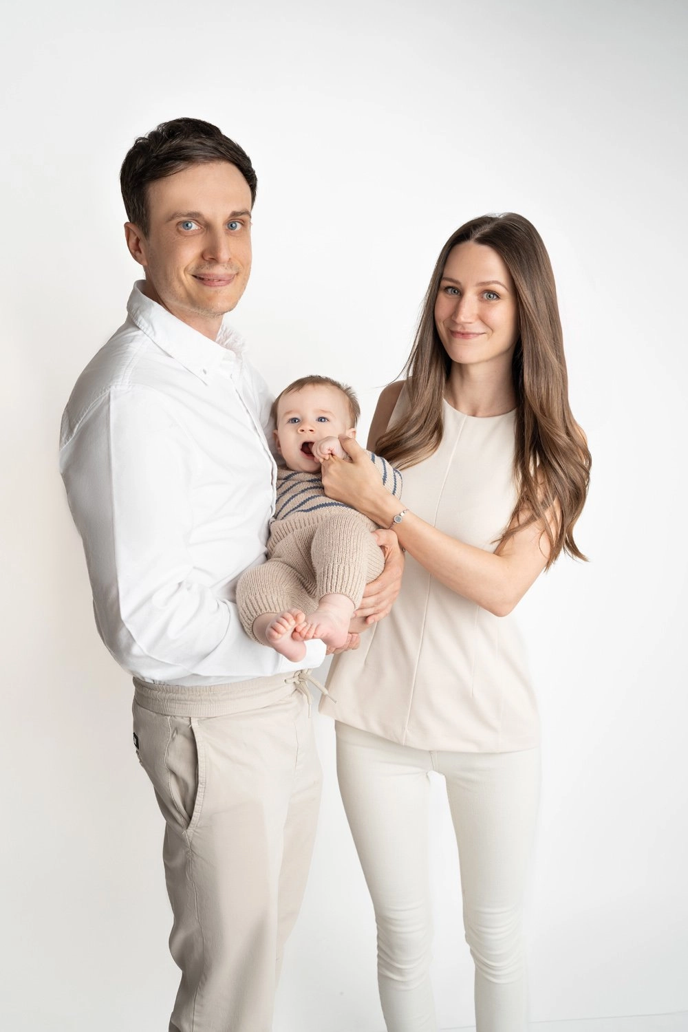 Zwei Erwachsene in heller Kleidung halten ihr Baby vor weißem Hintergrund.