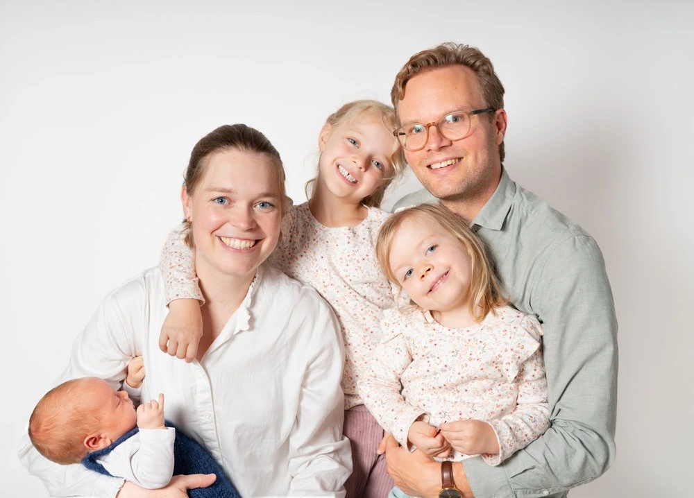 Zwei Erwachsene kuscheln mit drei Kindern vor einem hellen Hintergrund.