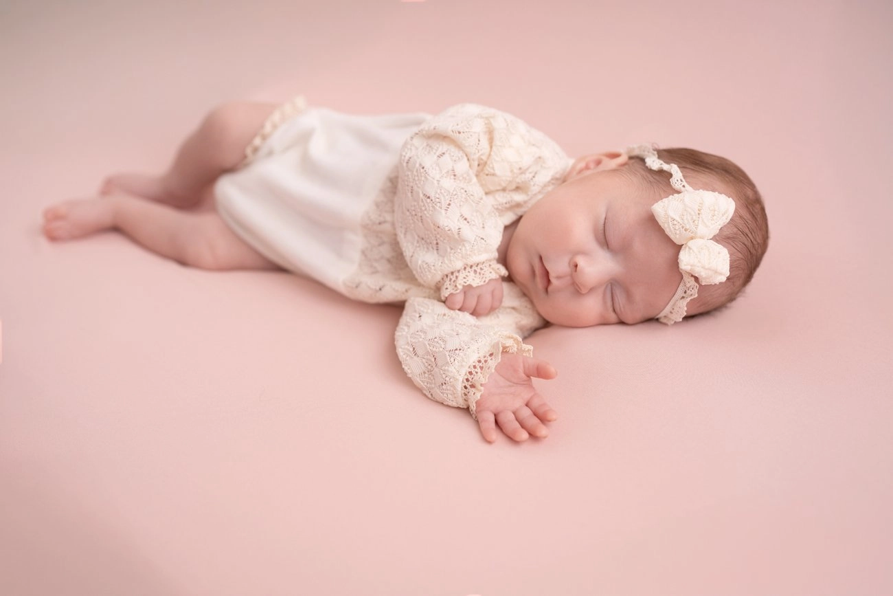 Ein schlafendes Baby in heller Spitzenkleidung liegt auf rosa Stoff.
