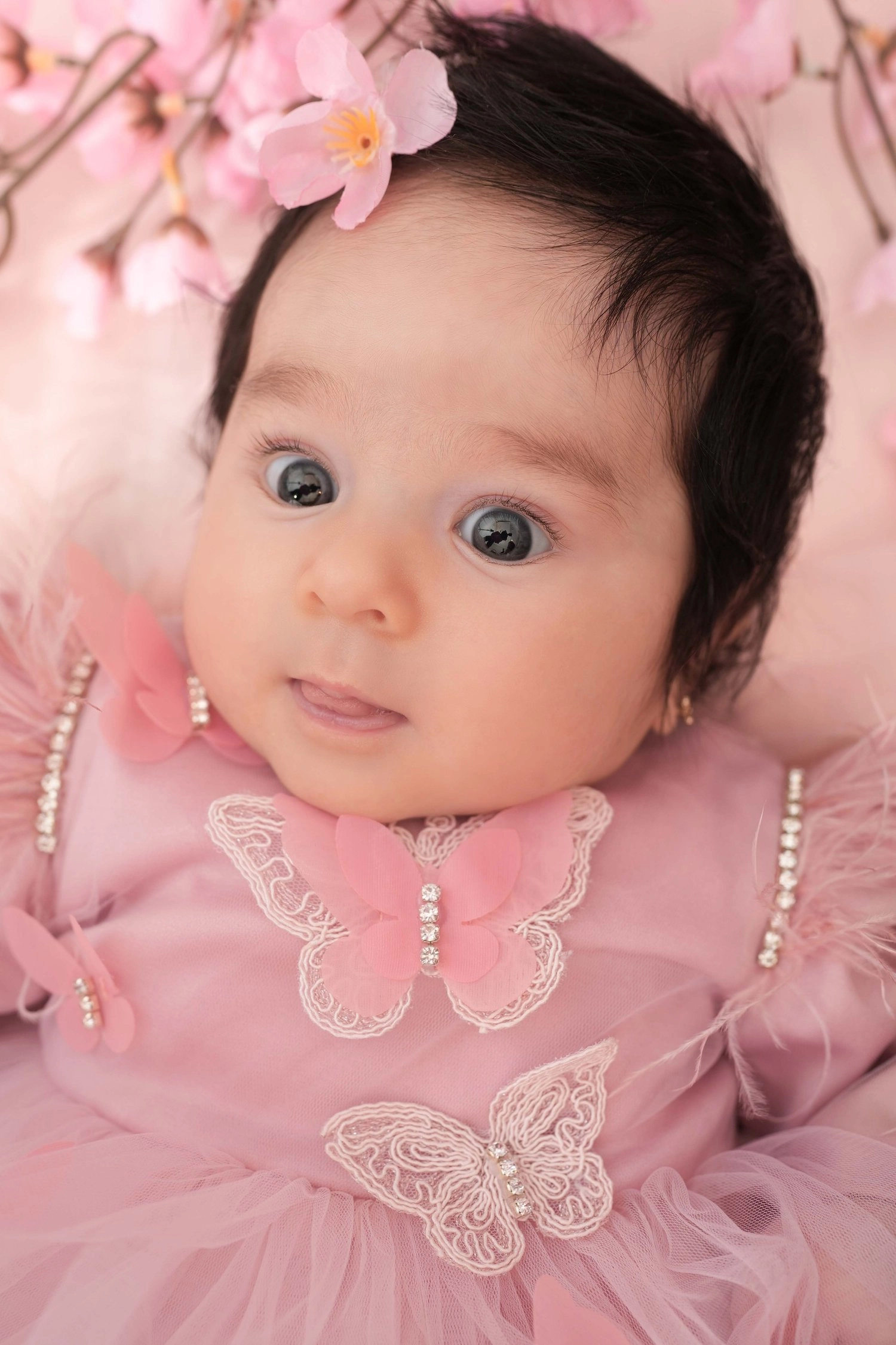 Ein waches Baby im rosa Kleid blickt unter rosa Blüten hervor.