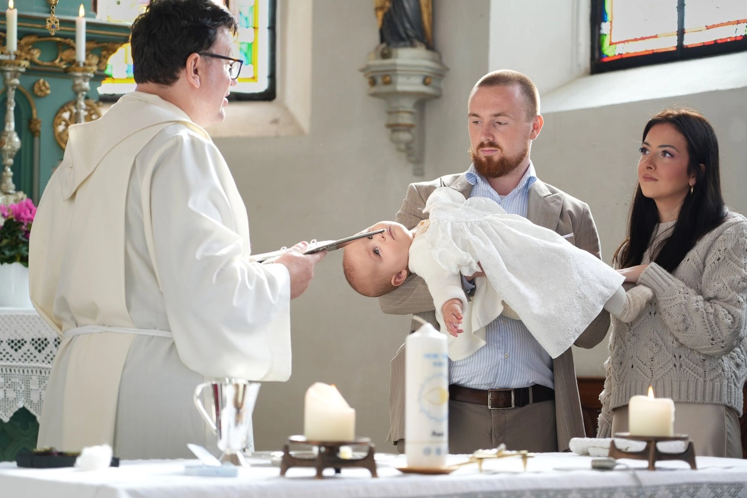 Ein Pfarrer steht einer Familie mit Baby am Taufbecken gegenüber.