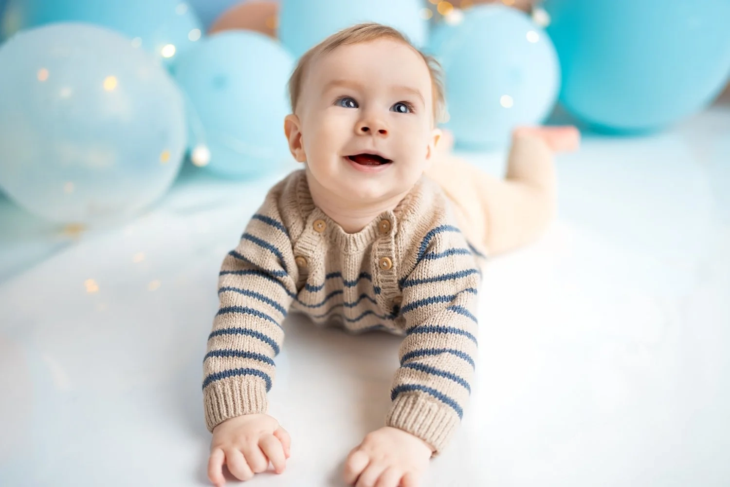 Ein lachendes Baby liegt im Streifenpullover vor hellblauen Ballons.