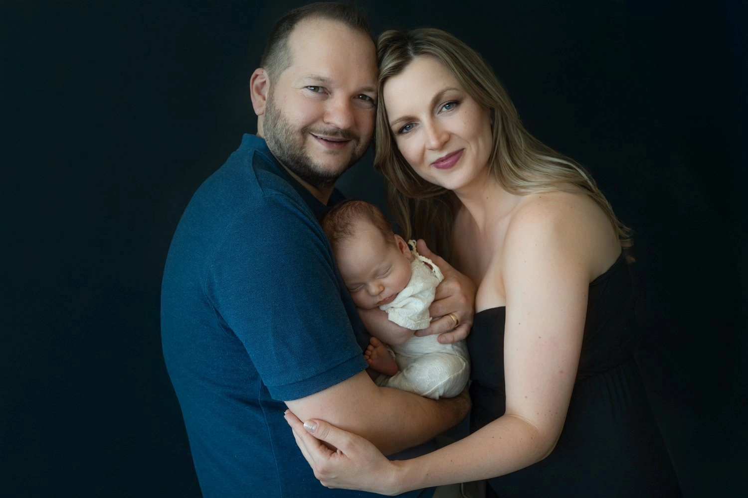 Zwei Erwachsene halten ein schlafendes Baby vor einem dunklen Hintergrund.