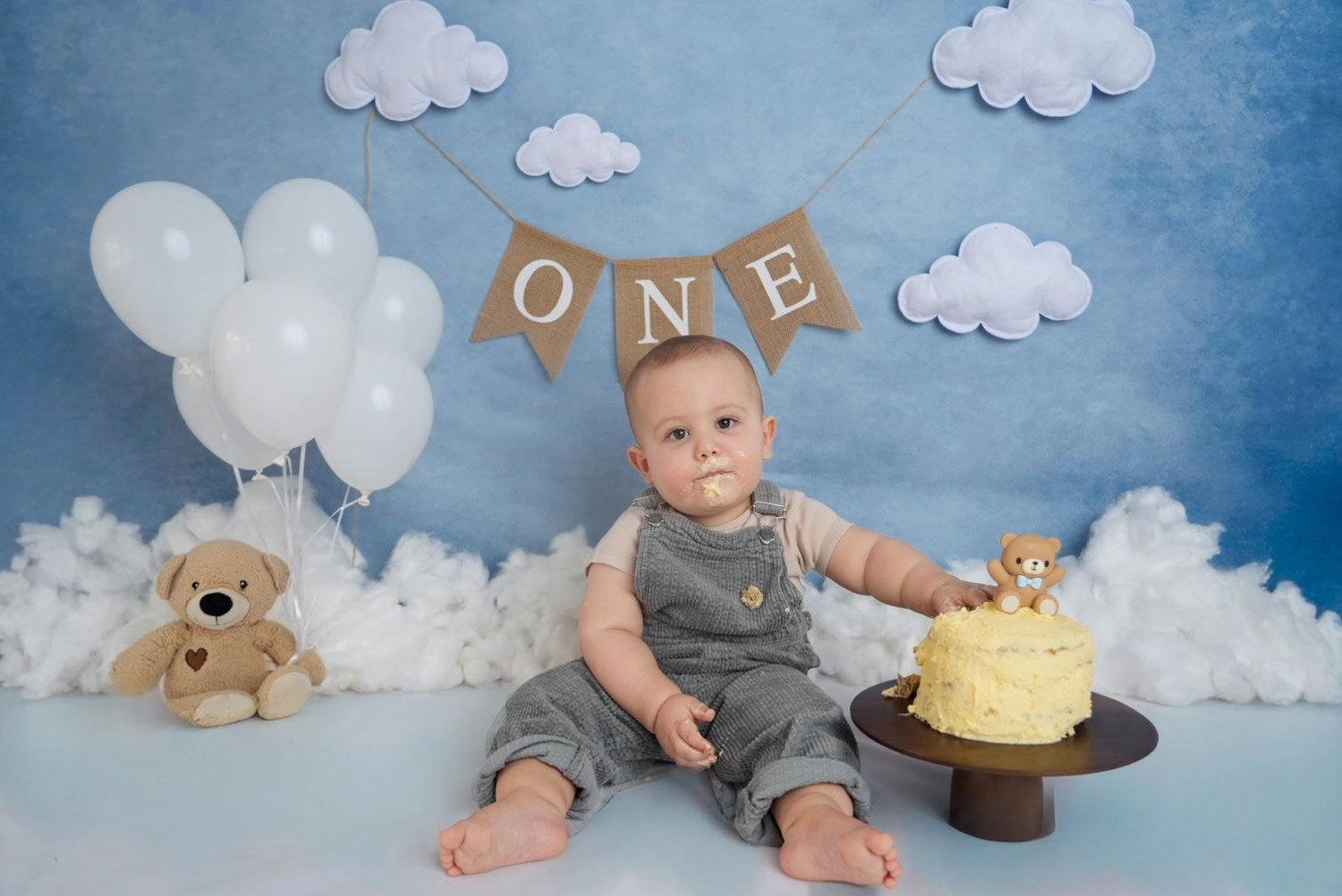 Ein Baby sitzt neben Torte und Teddybär vor einer blauen Wolkenkulisse.