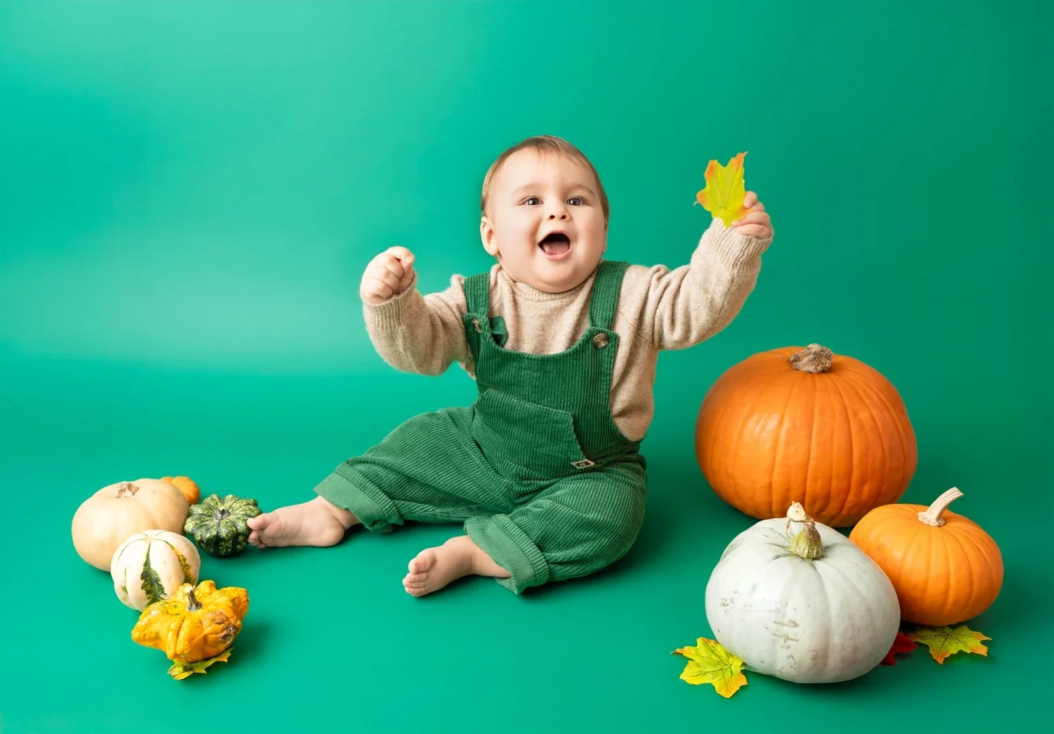 Ein Baby im grünen Outfit hebt zwischen Kürbissen ein Herbstblatt.