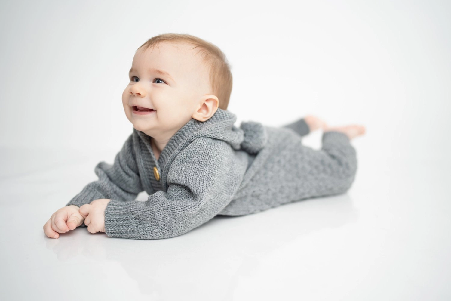 Ein Baby im grauen Strickpullover liegt lächelnd auf hellem Untergrund.