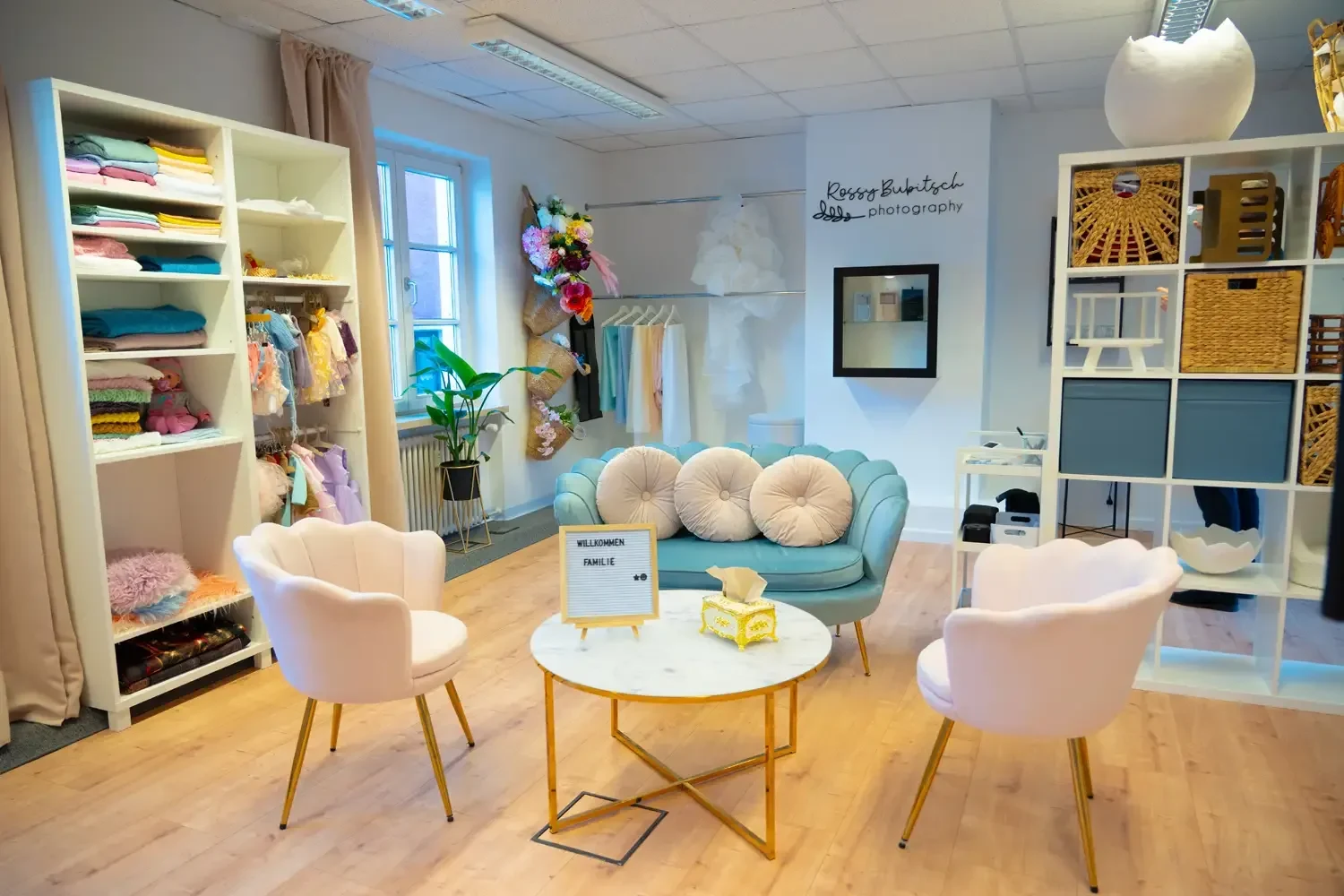 Ein heller Studioraum zeigt Sofa, Sessel, Tisch und Kleidung im Regal.