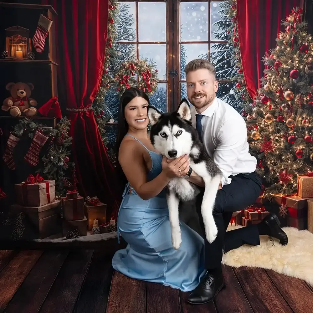 Ein Paar hält seinen Husky zwischen Weihnachtsbaum und festlich geschmücktem Fenster.