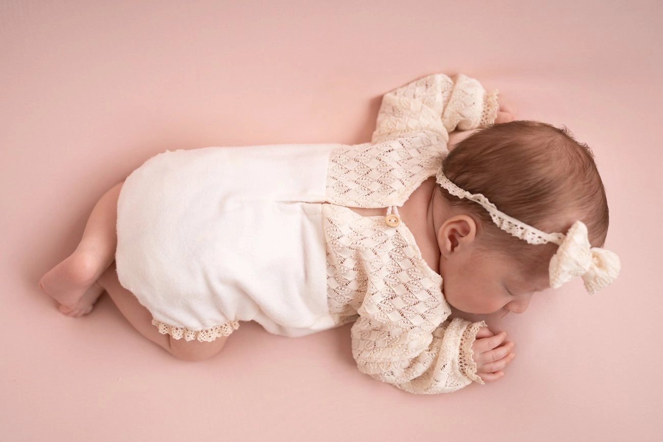 Ein schlafendes Baby mit Schleifenhaarband liegt auf rosa Stoff, von oben gesehen.