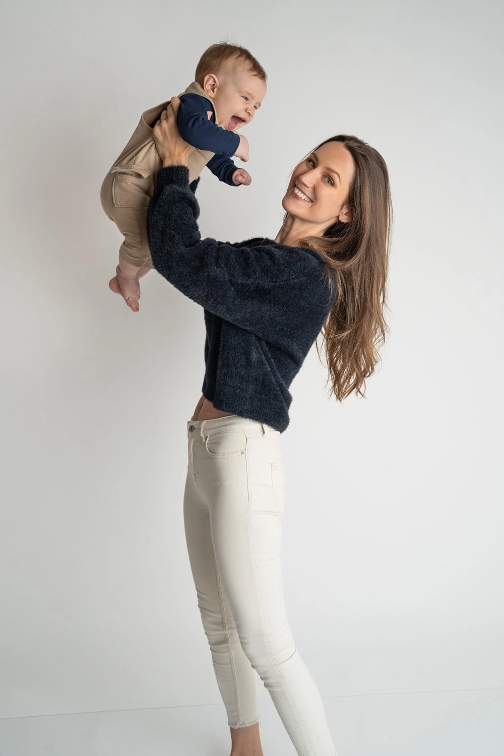 Eine Frau im dunklen Pullover hebt ein Baby lächelnd hoch.