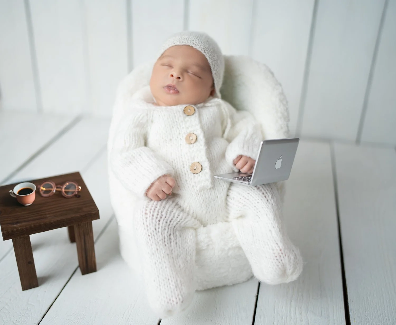 Ein schlafendes Baby im weißen Strickanzug liegt auf einem kleinen Sessel.
