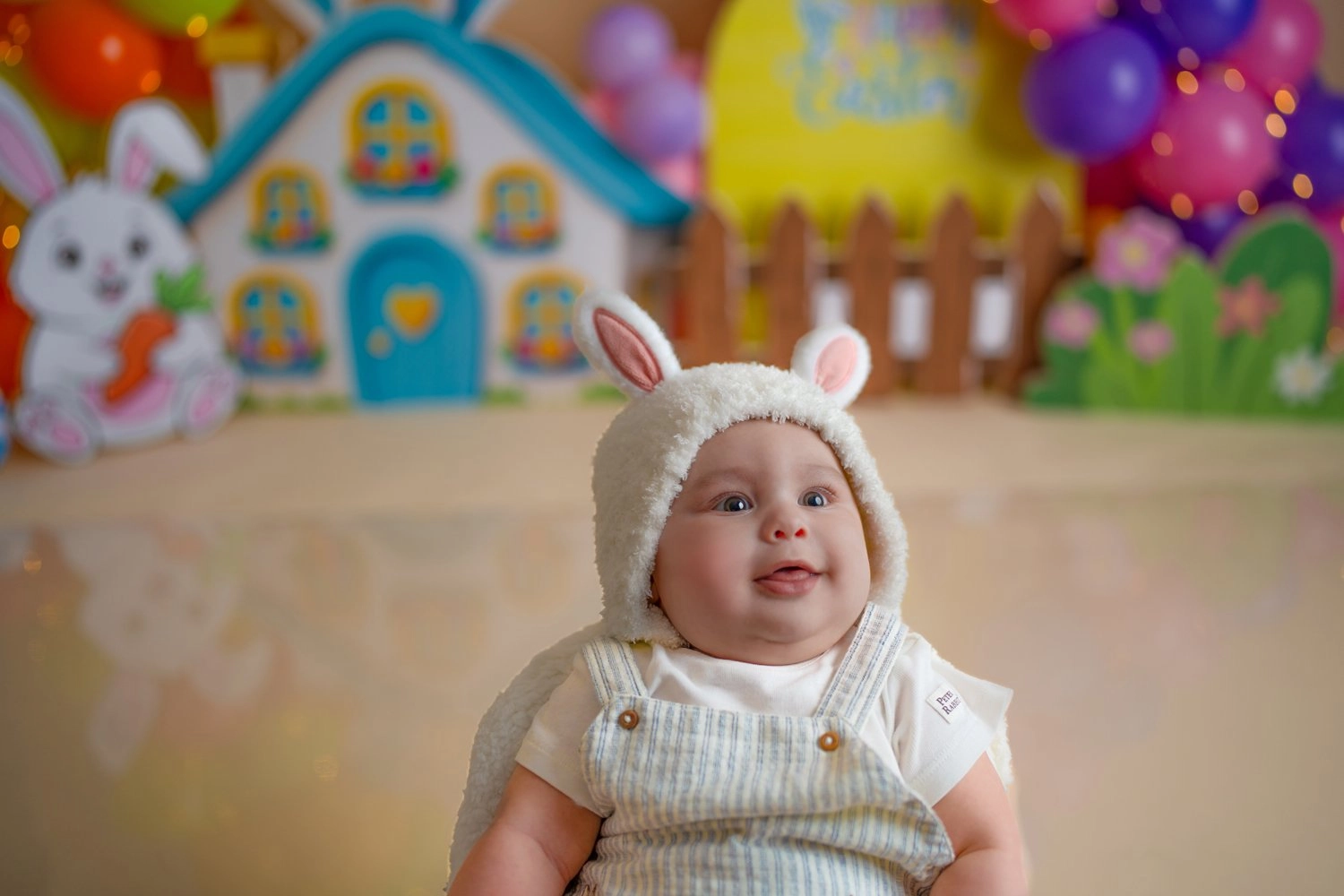 Ein Baby mit gestrickten Hasenohren sitzt vor einer bunten Osterkulisse.