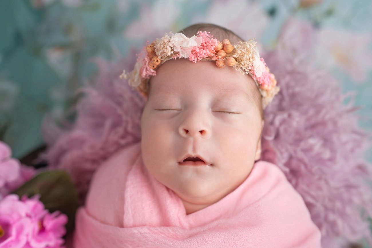 Ein schlafendes Baby mit Blumenkranz ist in rosa Stoff eingewickelt.